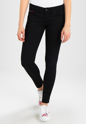 Frau trägt schwarze Slim-Fit-Jeans und weiße Sneakers mit roter Blumenstickerei, steht vor einem schlichten Hintergrund.