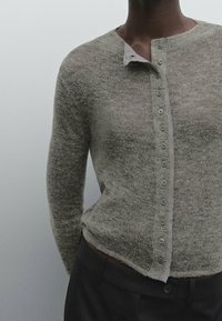 Lähikuva henkilöstä, joka pukeutuu tekstuurilla varustettuun harmaaseen napitettavaan neuleeseen ja tummaan housuun yksinkertaista harmaata taustaa vasten.