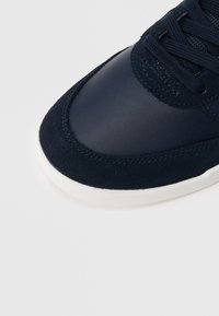 Detailní záběr na špičku námořnicky modré tenisky se semišovými a koženými texturami, vybavené stejnobarevnými tkaničkami a bílou gumovou podrážkou.