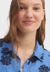 Chemise à col bleu arborant un motif floral noir, fabriquée en tissu lisse avec une coupe décontractée. Design à boutons, boutons visibles.