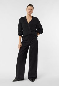 Kobieta stojąca z ręką w kieszeni, ubrana w czarny, fakturowany sweter o kopertowym kroju, szerokie czarne spodnie z subtelnym połyskiem oraz czarne buty w szpic.