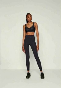 Egy nő magabiztosan áll, fekete sportmelltartót, magas derekú leggingset és fekete tornacipőt viselve, egy sima világos háttér előtt.