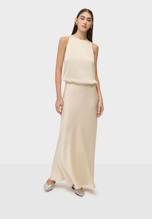 Kobieta stojąca w bezrękawowej beżowej sukni maxi z marszczoną talią i srebrnymi szpilkami z ostrym noskiem na jednolitym tle.