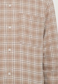 Barna kockás flaneling fehér vonalakkal, gombokkal, mellzsebbel és puha textúrával. A minta sötét és világos barna négyzetek váltakozásából áll.