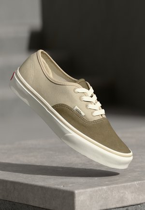 Low-Top Vans Sneaker mit beigem und olivgrünem Wildleder-Obermaterial, weißen Schnürsenkeln und weißer Gummisohle, schweben über einer Steinoberfläche.