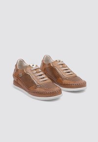 Zapatillas de cuero marrón con diseño perforado, suela blanca plana y costuras decorativas a lo largo de los bordes. Cuenta con cordones elásticos para facilitar su uso.