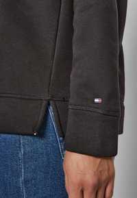 Gros plan d'une personne portant un haut à manches longues foncé avec un petit logo rouge, blanc et bleu près du poignet, et un jean en denim bleu.