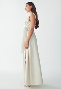 Robe longue couleur crème en tissu texturé avec un détail de nœud sur le côté. Elle présente un design sans manches et descend jusqu'au sol.