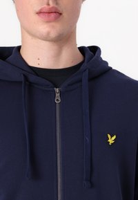 Sudadera con cremallera de color navy, con cordones y un logo bordado amarillo en el pecho. Hecha de una tela suave con puños de canalé y un diseño simple.