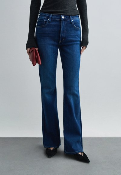 Persoon die donkerblauwe uitlopende jeans, zwarte puntige schoenen, een zwart shirt met lange mouwen draagt en een opgevouwen rode clutch vasthoudt tegen een effen achtergrond.