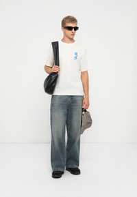 White cotton t-shirt with blue graphic, loose-fitting blue denim jeans, black bag, black shoes, and gray cap. Sunglasses complete the look.