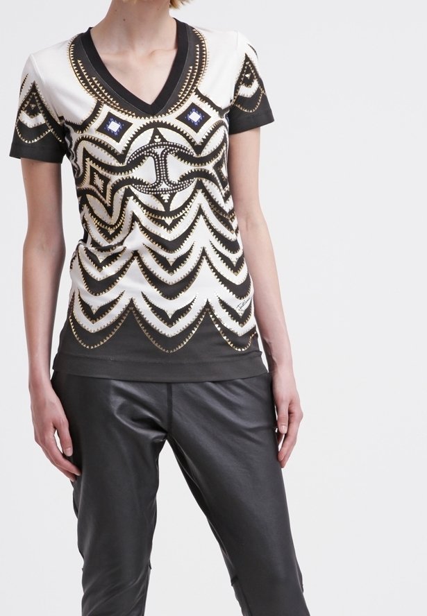 Černý a bílý top s krátkým rukávem a výstřihem do V, zdobený zlatými a modrými geometrickými vzory; kombinovaný s černými kalhotami z umělé kůže.