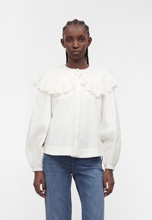 Frau mit geflochtenem Haar, die eine weiße Bluse mit großem Lochspitzenkragen und Puffärmeln trägt, kombiniert mit blauen Jeans, vor einem weißen Hintergrund.