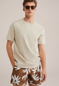 Beige bavlnené tričko s krátkym rukávom v kombinácii s hnedými plavkovými šortkami s veľkými bielymi kvetinovými vzormi. Model nosí slnečné okuliare.