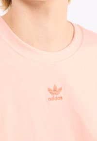 Hellrosa Baumwoll-T-Shirt mit Rundhalsausschnitt und kleinem, geprägtem adidas-Logo in der Mitte. Glatte Textur und lässiges Design.