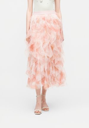 Fließender Midirock aus mehreren Schichten hellrosa und pfirsichfarbenem Blütentüll, kombiniert mit nudefarbenen Sandalen mit Knöchelriemen, vor weißem Hintergrund.