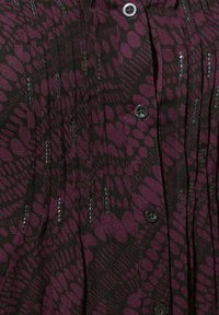 Blusa viola e nera con un design a pieghe, caratterizzata da motivi circolari, dettagli in paillettes e bottoni neri lungo il fronte.