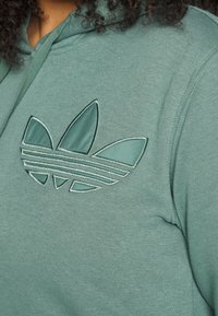 Grön hoodie med en präglad logotyp som har tre blad och horisontella ränder, tillverkad av mjukt tyg med en slät textur.