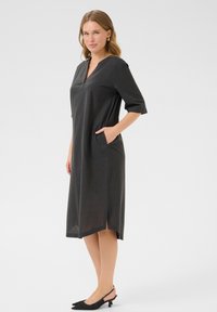 Robe noire mi-longue avec un décolleté en V, des manches trois-quarts, des poches latérales et des fentes sur les côtés. Texture douce avec une finition lisse.