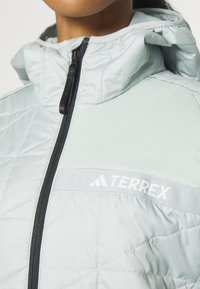 Veste isolante gris clair avec une texture matelassée, dotée d'une fermeture éclair noire et d'une capuche. Marquage "TERREX" en blanc sur le devant.