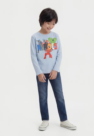 Niño con camiseta de manga larga azul claro con estampado colorido de superhéroes y la palabra "HEROES", pantalones vaqueros azules y zapatillas blancas, sonriendo hacia abajo.