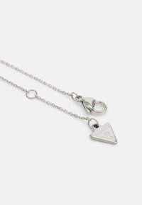 Collier en chaîne argentée avec un pendentif triangulaire arborant un logo "GUESS" gravé, sécurisé par un fermoir à ressort et des détails en anneau circulaire.