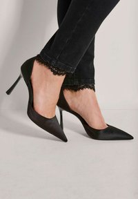 Vrouw in zwart puntig stilettohakschoen en zwarte broek met kantafwerking bij de enkels tegen een effen lichte achtergrond.