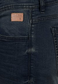 Tmavě modré džíny z denimu zblízka, ukazující zadní kapsy a malou hnědou koženou nášivku se textem "BLEND Místní výrobce džín od roku 1993".