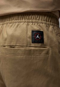 Brune stof joggere med en strækbar talje, der har en baglomme og et sort mærke med et rødt Jumpman-logo.