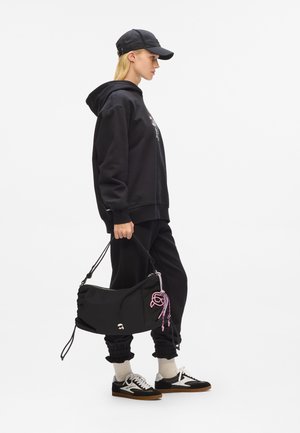 Schwarze Schultertasche mit matter Textur, ausgestattet mit pinken Akzenten und einem Kordelzugverschluss. Das Modell trägt einen schwarzen Hoodie und eine Sweatpants, kombiniert mit Sneakers.