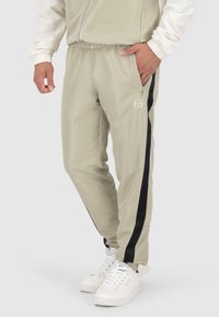 Helle beige Jogginghose mit schmalem Schnitt, schwarzen Seitenstreifen, elastischem Bund und funktionalen Taschen, kombiniert mit weißen Sneakers.