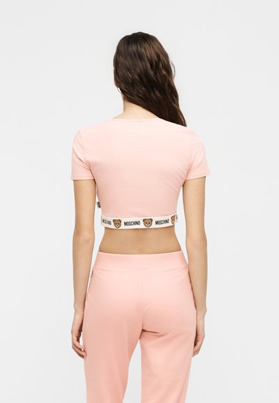 Γυναίκα με μακριά κυματιστά μαλλιά που φοράει ανοιχτό ροζ κοντό τοπ και ταιριαστό παντελόνι, με λωρίδα στη μέση με το λογότυπο αρκουδάκι Moschino.