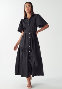 Čierne šaty na gombíky s opaskom v páse, krátkymi pufovými rukávmi a vrstvenou rozšírenou sukňou. Vyrobené z ľahkej tkaniny s hladkou textúrou.