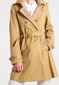 Trench coat beige con bottoni doppi, vita con cintura e ampio colletto. Presenta maniche lunghe con polsini abbottonati e linee pulite.