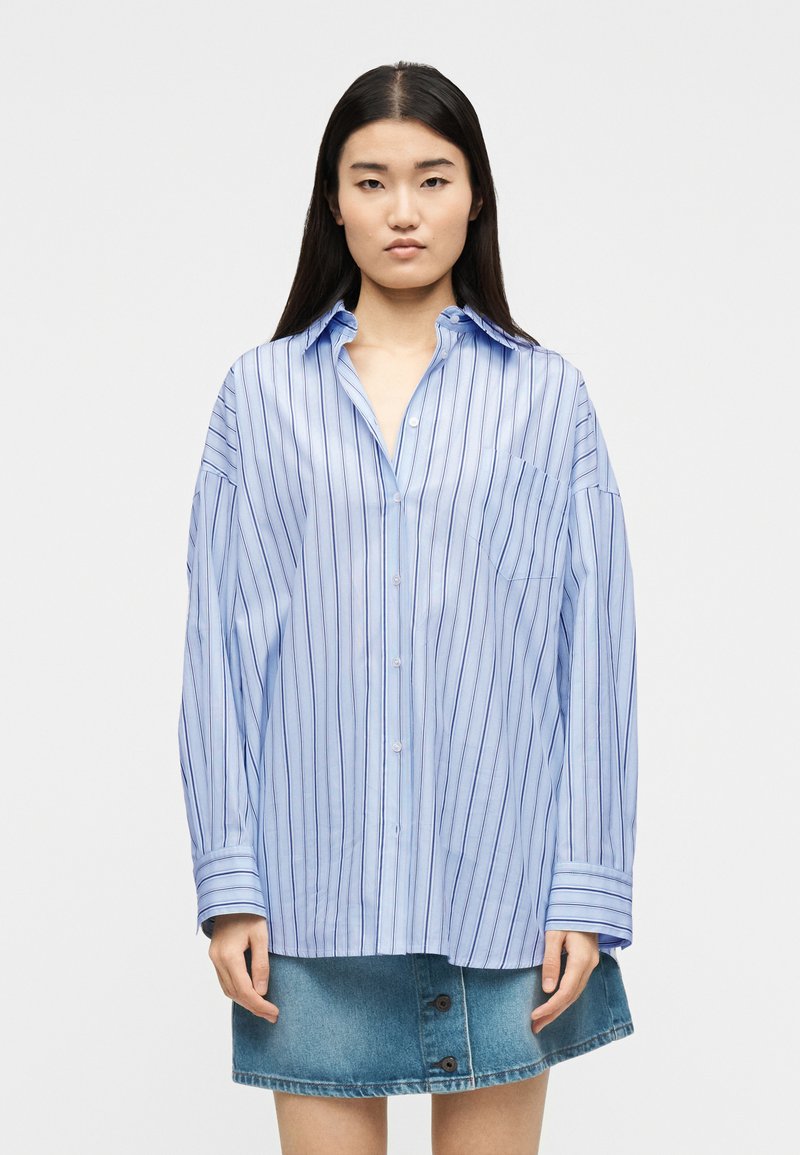 Νεαρή γυναίκα με μια υπερμεγέθη γαλάζια ριγέ πουκάμισο με κουμπιά και μια μπλε τζιν φούστα με κουμπιά μπροστά, στέκεται μπροστά σε απλό λευκό φόντο.