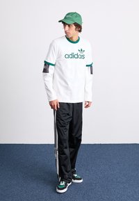 Fehér, hosszú ujjú ing zöld díszítéssel és "adidas" logóval, fekete sportnadrággal, fehér oldalsávokkal, valamint zöld sportcipővel párosítva.