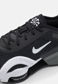 Černá sportovní bota s prodyšným síťovaným svrškem, texturovaným povrchem, bílým logem Nike a viditelným odpružením v podrážce. Šňůrky jsou černé, logo bílé.