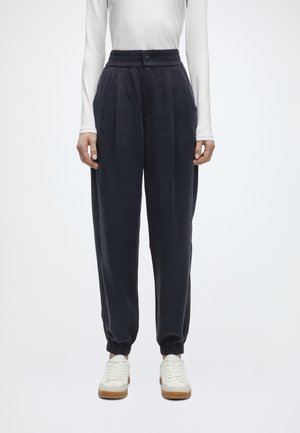 Person trägt hochgeschnittene, lockere schwarze Jogginghosen mit elastischen Bündchen und weißen Sneakers, kombiniert mit einem eng anliegenden weißen Langarmshirt.