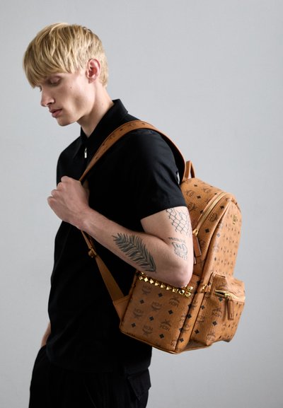 Sac à dos en cuir tan avec des motifs gaufrés, des clous dorés et des fermetures éclair. Présente une finition texturée et des bretelles réglables.