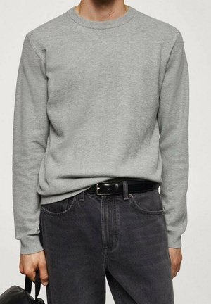 Person trägt einen hellgrauen Pullover mit langen Ärmeln, schwarzen Gürtel, dunkelgraue Jeans und hält eine schwarze Tasche an der Seite.