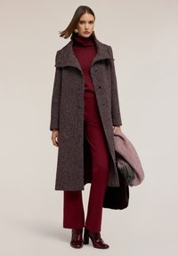 Cappotto lungo, bordeaux e crema con motivo, dotato di un colletto alto, abbinato a un dolcevita bordeaux, pantaloni coordinati e stivali con tacco alto.
