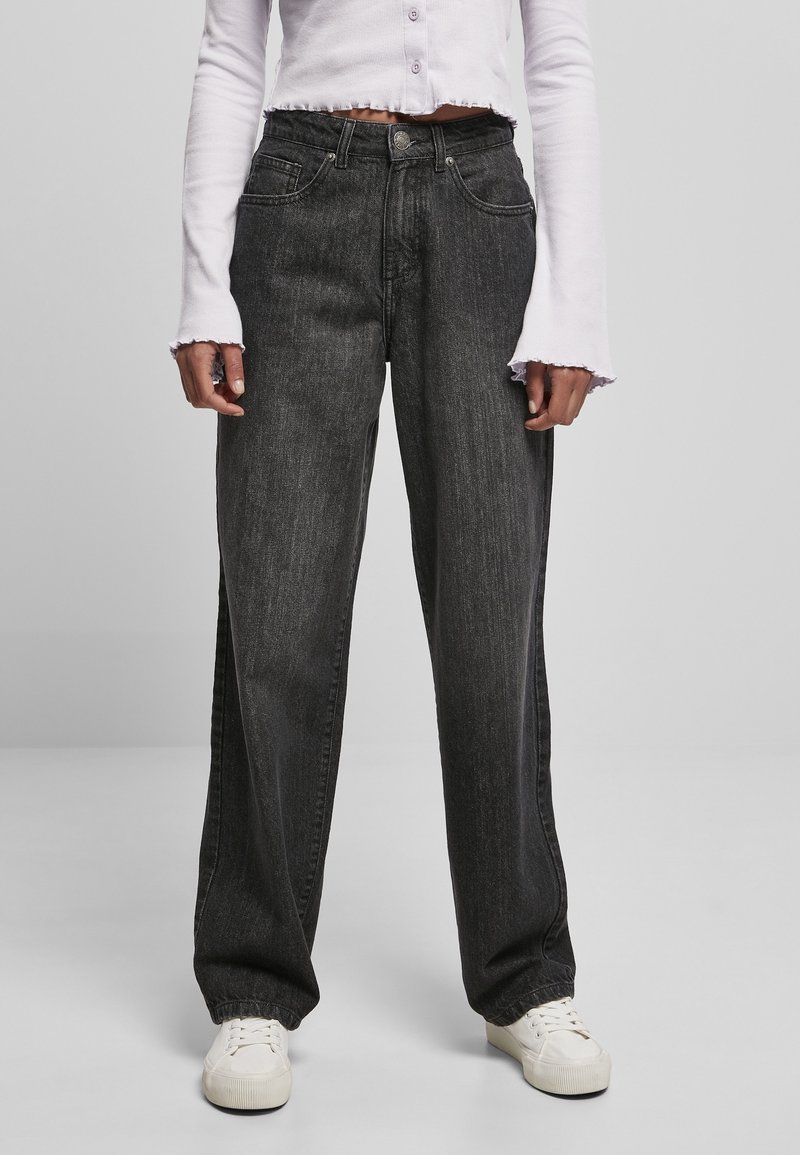 Jeans negros de cintura alta, diseño de pierna ancha, textura desgastada, estilo de cinco bolsillos, combinados con zapatillas blancas y una blusa de manga larga recortada.