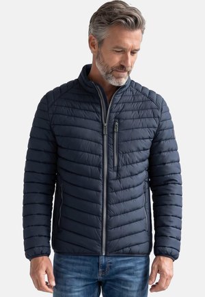 Uomo con capelli grigi e barba, indossa una giacca trapuntata blu navy con cerniera e jeans blu, guarda verso il basso con le mani rilassate lungo i fianchi.