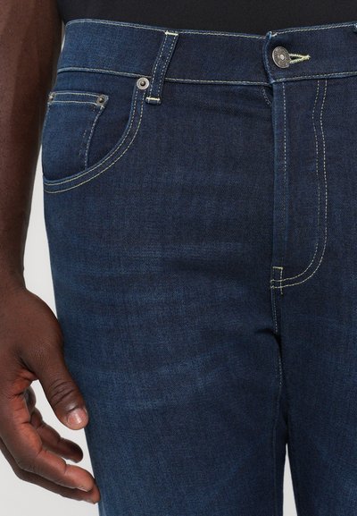 Jeans din denim albastru închis cu cusături galbene, având un buzunar frontal și o închidere cu buton. Materialul are o textură netedă.