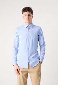 Camisa azul clara de manga longa com colarinho pontiagudo, frente com botões e textura suave, combinada com calças caqui.