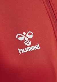 Rode stof met een wit logo met een bijontwerp boven het woord "hummel." Het materiaal lijkt glad met een getailleerde vorm.