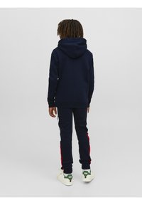 Sudadera azul marino con bolsillo canguro y capucha, combinada con pantalones jogger a juego que presentan rayas laterales rojas y acentos blancos. Se lleva con zapatillas blancas.