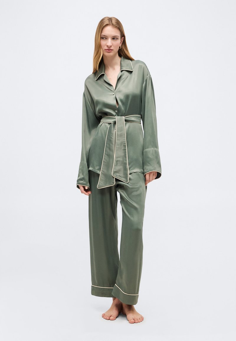 Γυναίκα όρθια, ξυπόλητη, φοράει χαλαρή, ελαιόχρωμη σατέν πιτζάμα με λευκή λεπτομέρεια, μακρυμάνικο τοπ με ζώνη και παντελόνι με φαρδιά μπατζάκια.