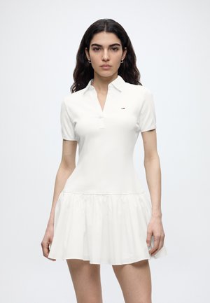 Kvinde med langt mørkt hår iført en hvid kortærmet kjole med krave, tætsiddende talje og udvidet skørt, stående mod en ensfarvet baggrund.