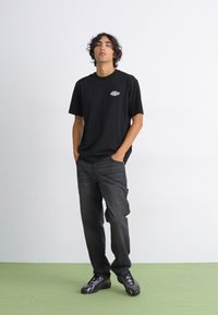 T-shirt noire à manches courtes avec un petit graphique sur la poitrine, associée à un jean gris foncé et des baskets noires, se tenant contre un fond uni.